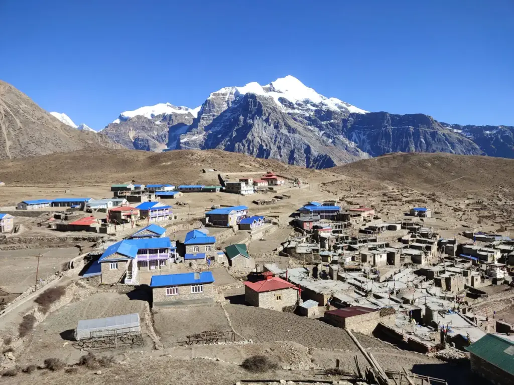 A high-altitude mountain village in the Nar Phu Valley featuring blue-roofed houses set against a massive snow-covered peak, ideal for monsoon treks in Nepal.