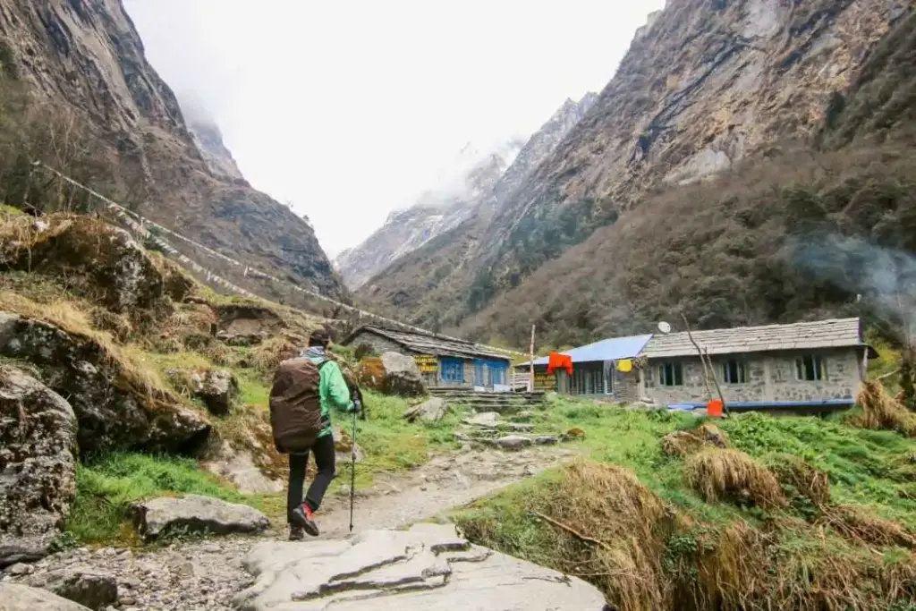 A trekker with a green jacket and backpack walks up a stone path toward rustic stone guest houses in a lush mountain valley, with steep cliffs and prayer flags in the background.